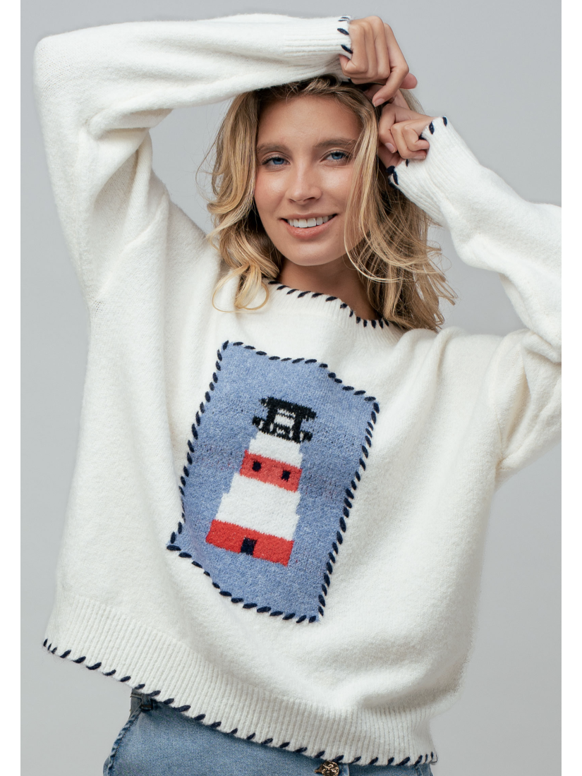 A woman wearing a cream knit sweater featuring a blue and white lighthouse design and blue whipstitch trim on the collar, cuffs, and bottom hem.