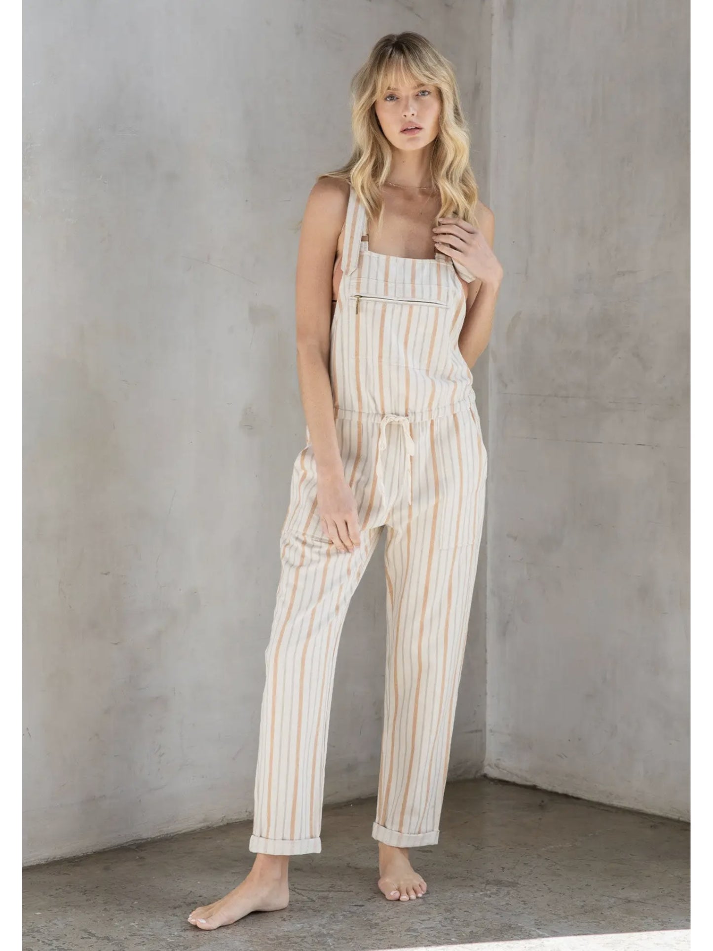 Woman in cream and tan striped linen-blend overalls with a drawstring waist and front zipper pocket, standing against a grey textured wall.