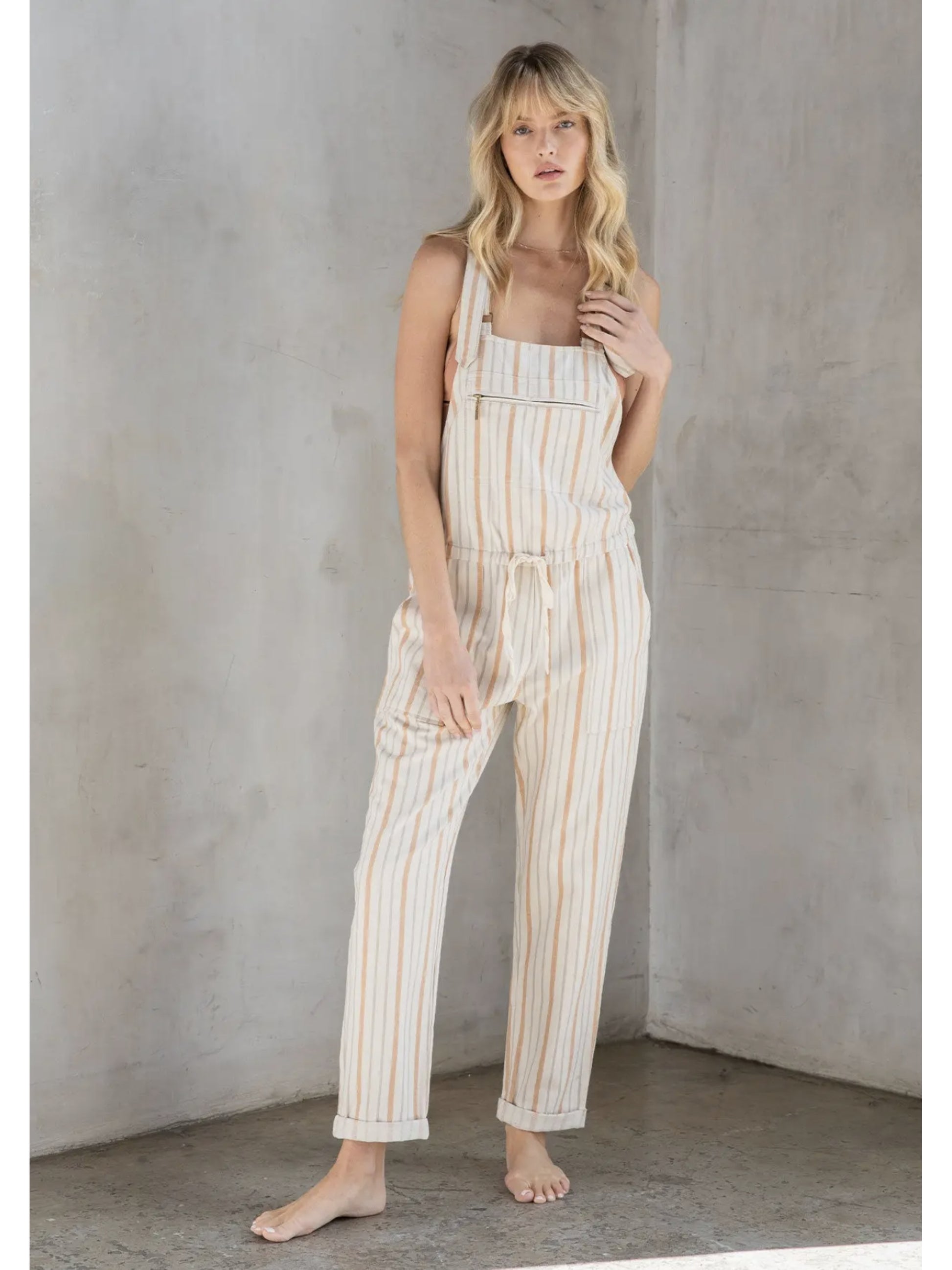 Woman in cream and tan striped linen-blend overalls with a drawstring waist and front zipper pocket, standing against a grey textured wall.