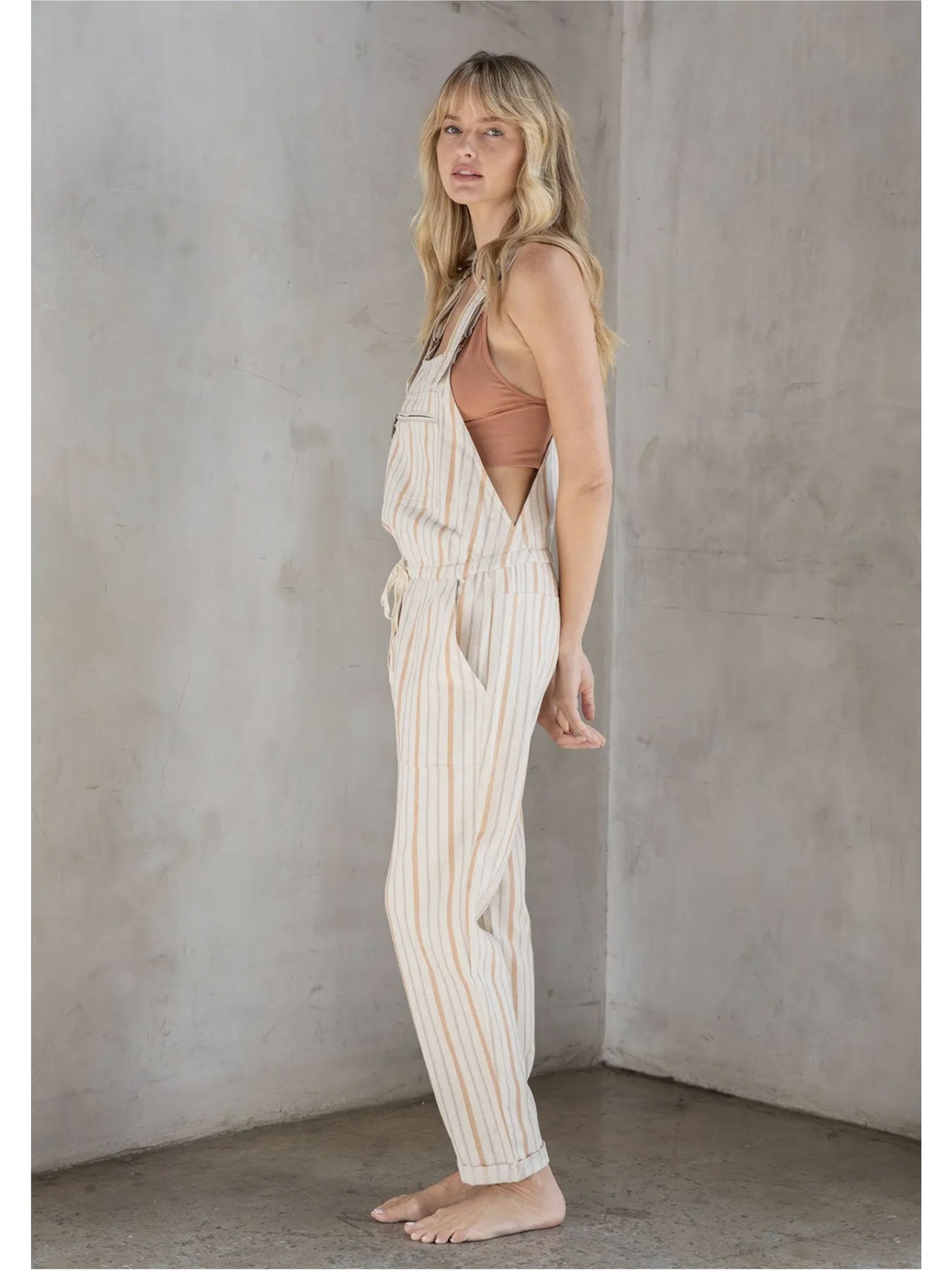 Woman in cream and tan striped linen-blend overalls with a drawstring waist and front zipper pocket, standing against a grey textured wall.