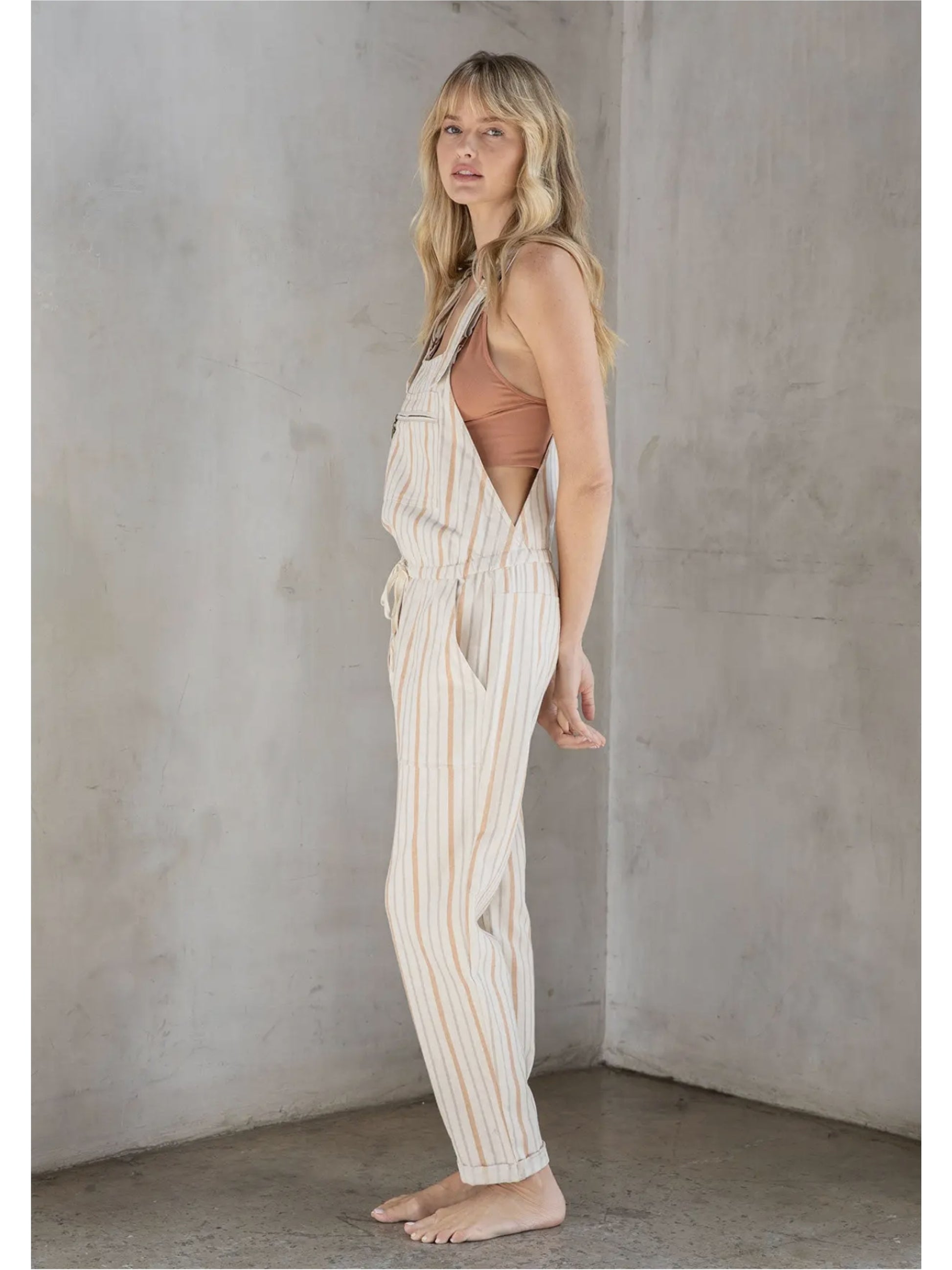 Woman in cream and tan striped linen-blend overalls with a drawstring waist and front zipper pocket, standing against a grey textured wall.