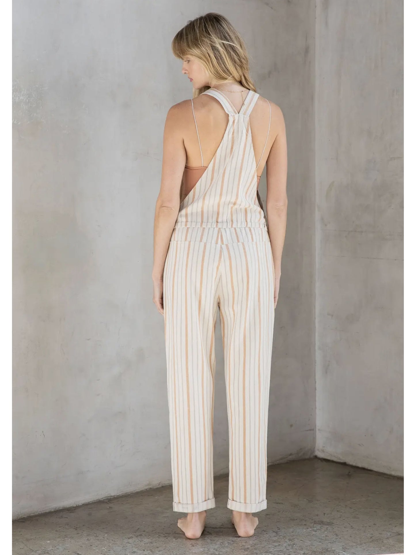 Woman in cream and tan striped linen-blend overalls with a drawstring waist and front zipper pocket, standing against a grey textured wall.