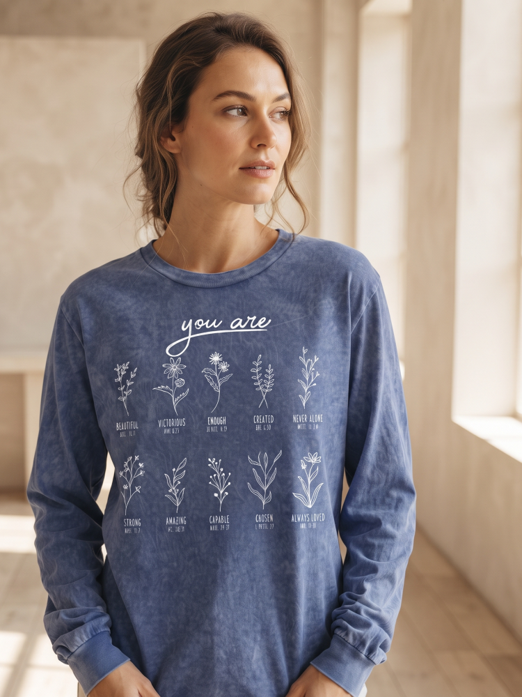 A woman wearing a blue long-sleeve crewneck sweatshirt with a white graphic print of various herbs and the words 'you are' on the front, set against a warm, sunlit indoor background.