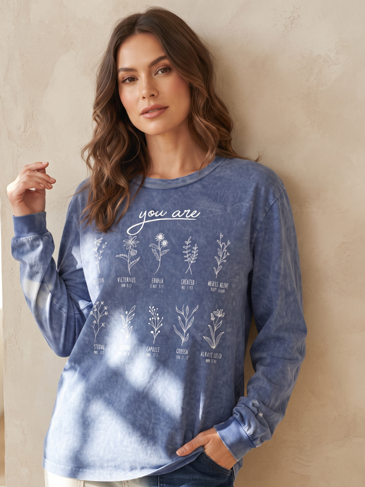 A woman wearing a blue long-sleeve graphic sweatshirt with white botanical illustrations and the text 'you are' on the front, standing against a textured beige wall.