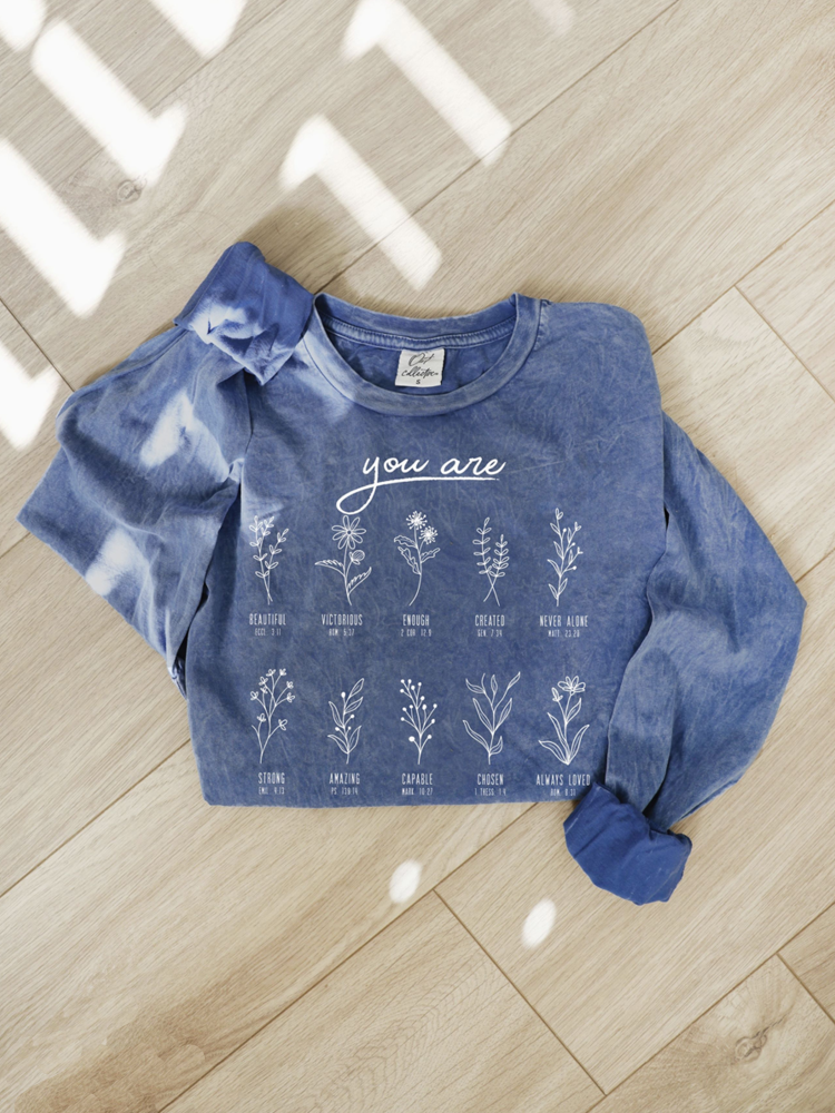 A blue tie-dye long-sleeve shirt with white botanical illustrations and the text 'you are' laid flat on a light wood floor with dappled sunlight.