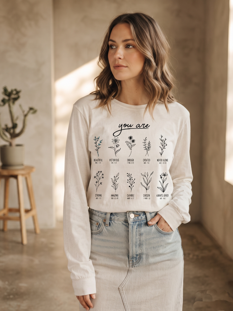 A woman wearing a white long-sleeve graphic t-shirt with black botanical illustrations and the text 'you are' and various affirmations underneath, paired with a light denim skirt in a warm, sunlit indoor setting.