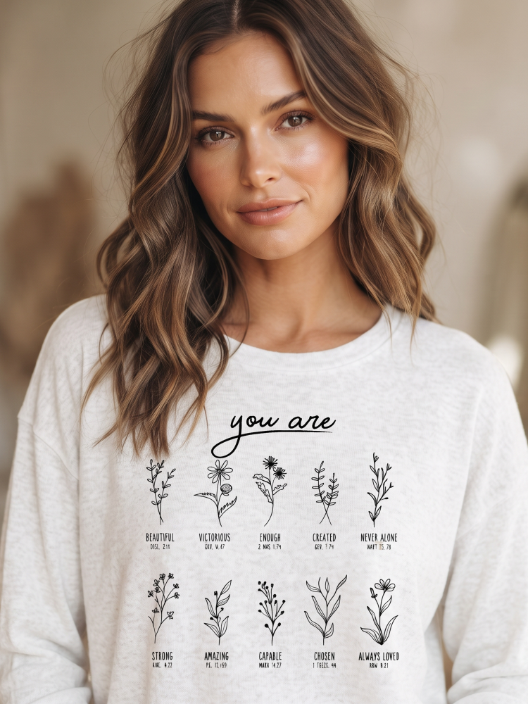 A woman with wavy brown hair wearing a light gray crewneck sweatshirt with a black graphic print of various flowers and the text 'you are'.