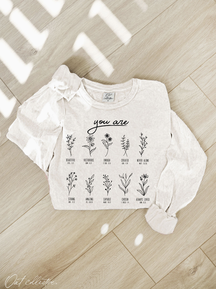 A white long-sleeve graphic sweatshirt with a bow on one sleeve, laid flat on a light wood floor. The front features the text 'you are' above a grid of illustrated plants with positive adjectives and small labels.